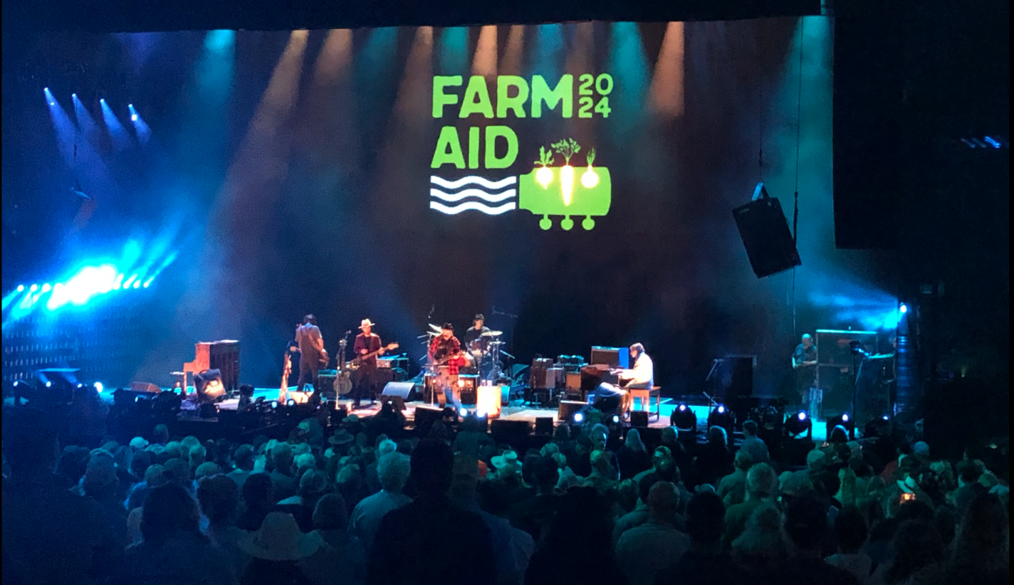 During the Farm Aid 2024 concert, musicians on stage perform in front of a large blue and green Farm Aid visual behind them. Colorful stage lights illuminate the audience.