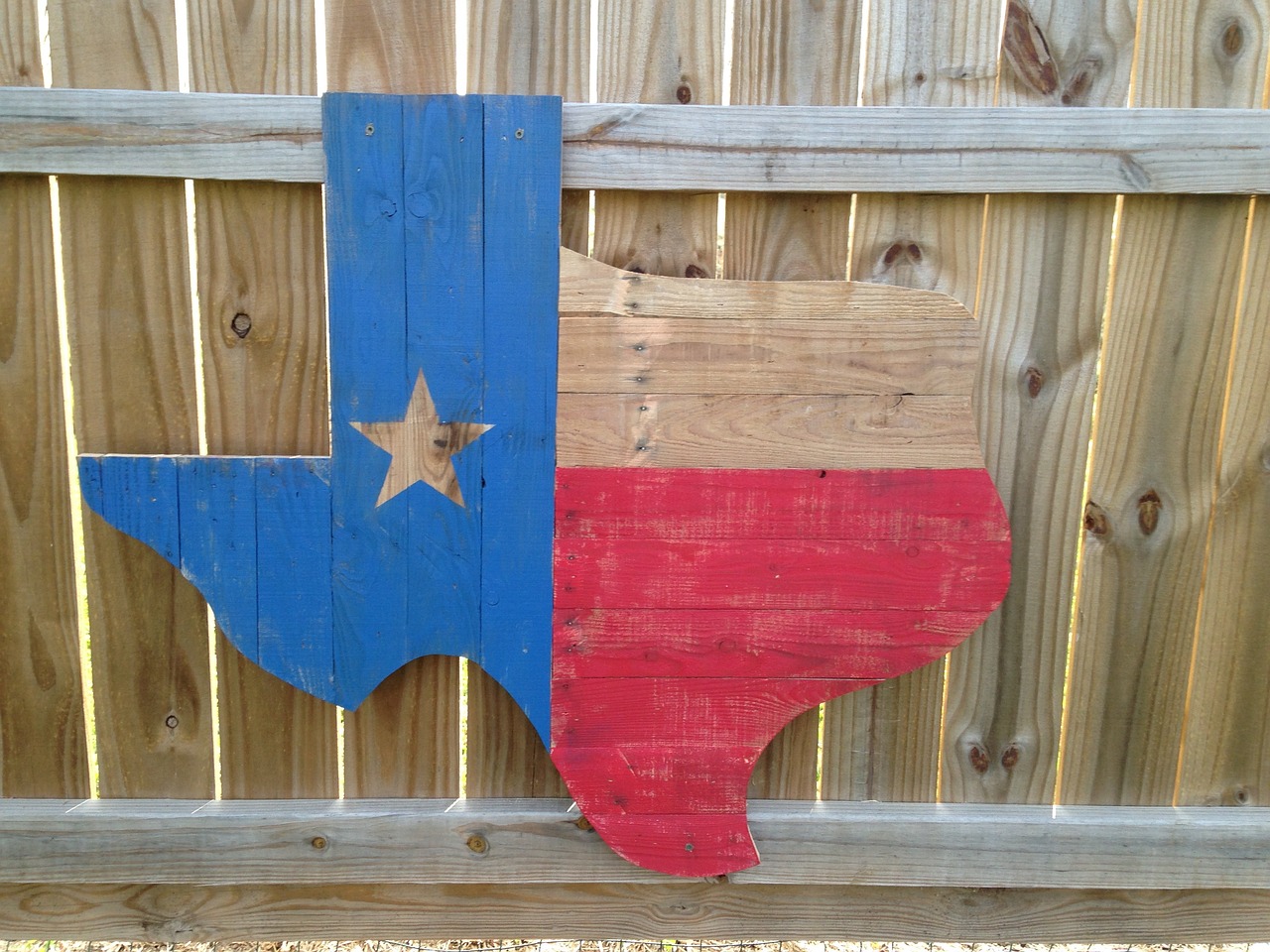 A wooden painted cutout in the shape of Texas and painted like the Texas flag sits atop a wooden fence.